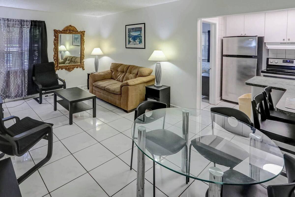 A modern living room with a sofa, a glass dining table, and a kitchen area with a refrigerator and stove.