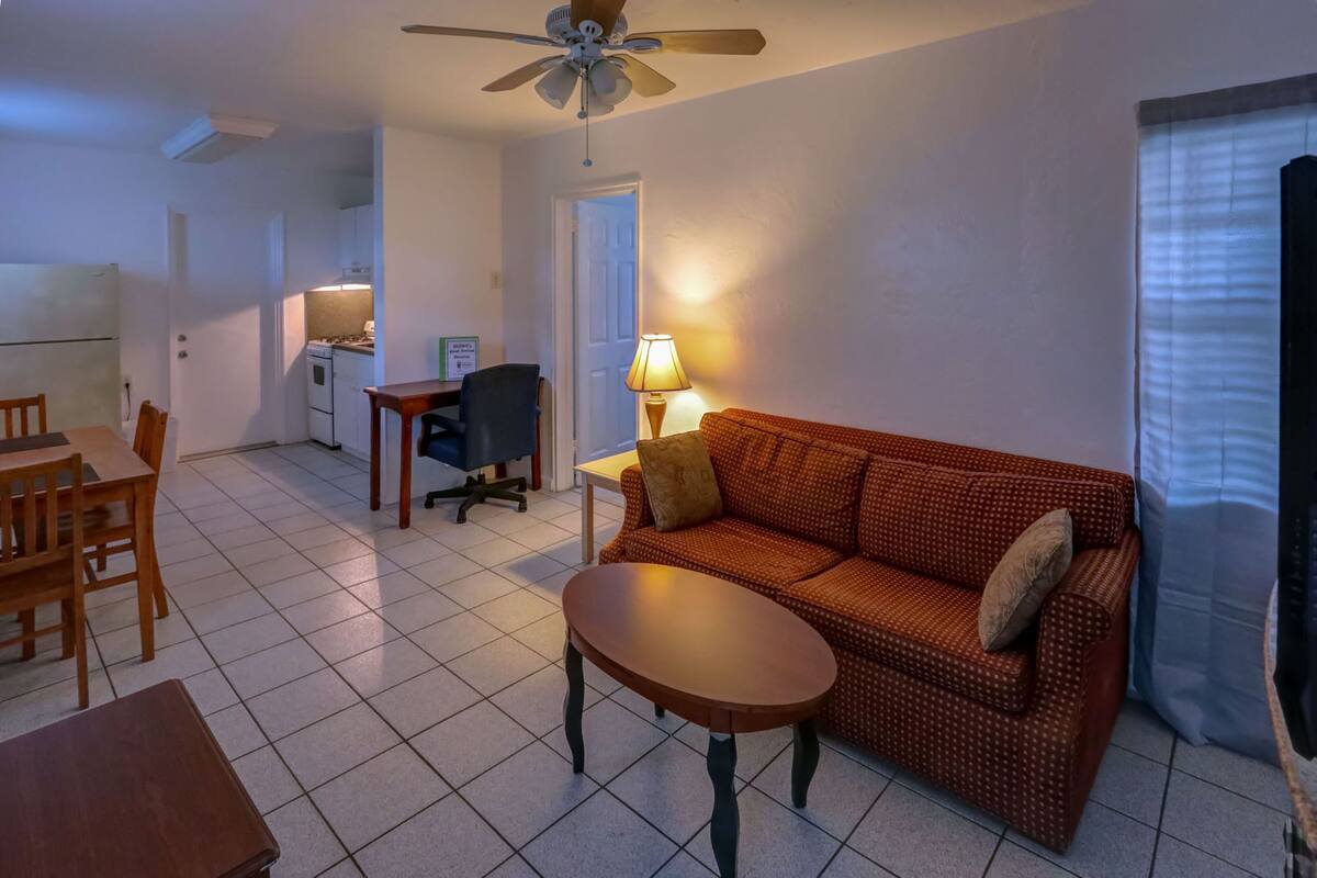 A living room with a sofa, a coffee table, and a dining area with a table and chairs. There is a kitchen area in the background.