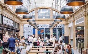Un vaste espace intérieur doté de hauts plafonds et de grandes fenêtres, comprenant un espace de détente central avec des tables et des chaises, ainsi qu’une devanture visible avec une enseigne indiquant un restaurant de pizzas.