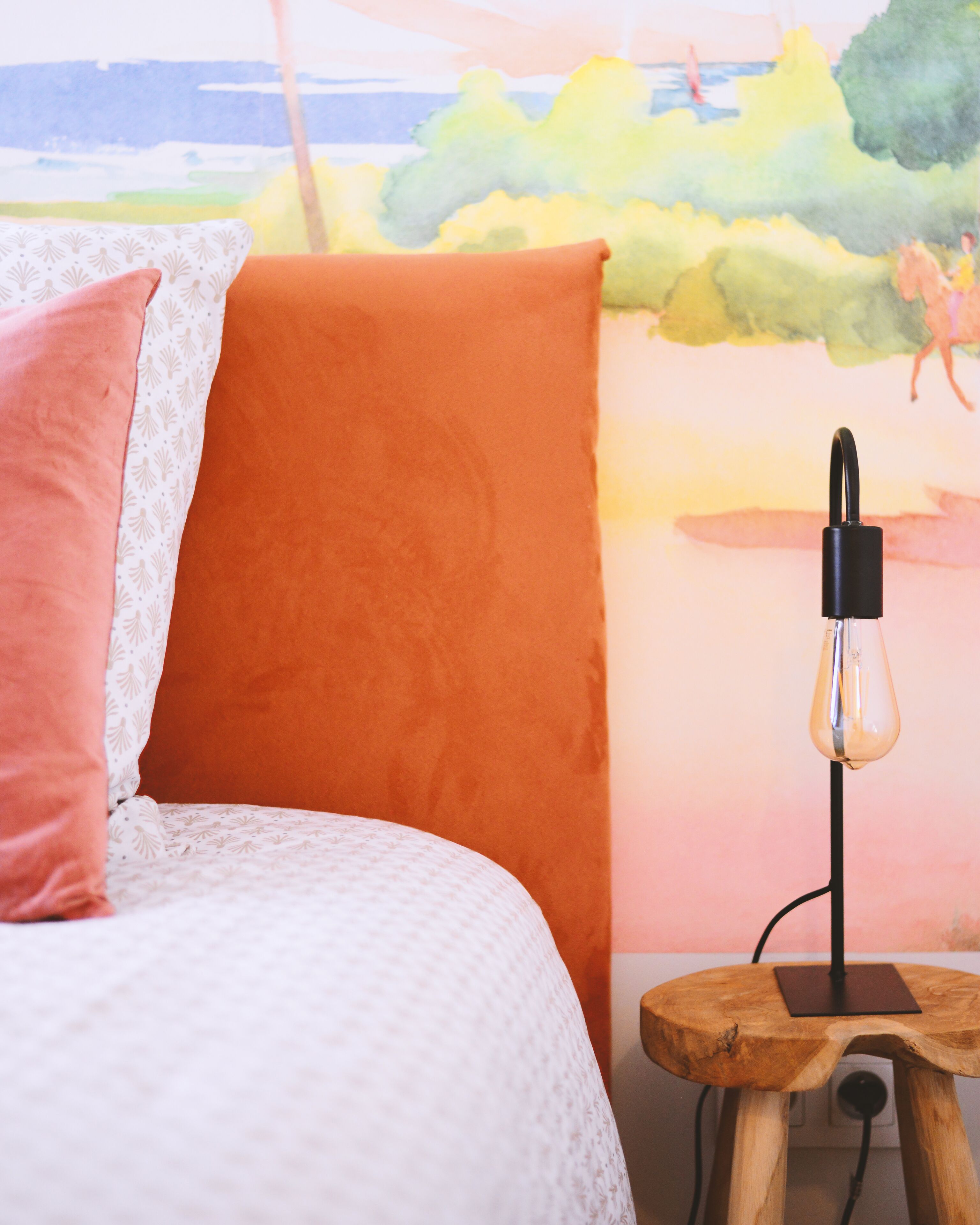 Een bed met een wit en oranje hoofdeinde, een roze kussen en een lamp op een houten nachtkastje.