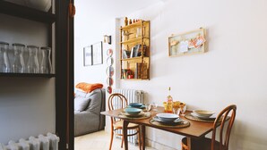 Un coin repas avec une table en bois dressée pour quatre personnes, entourée de chaises en bois. Une bibliothèque contenant divers objets est fixée au mur, et un radiateur est visible dans un coin.