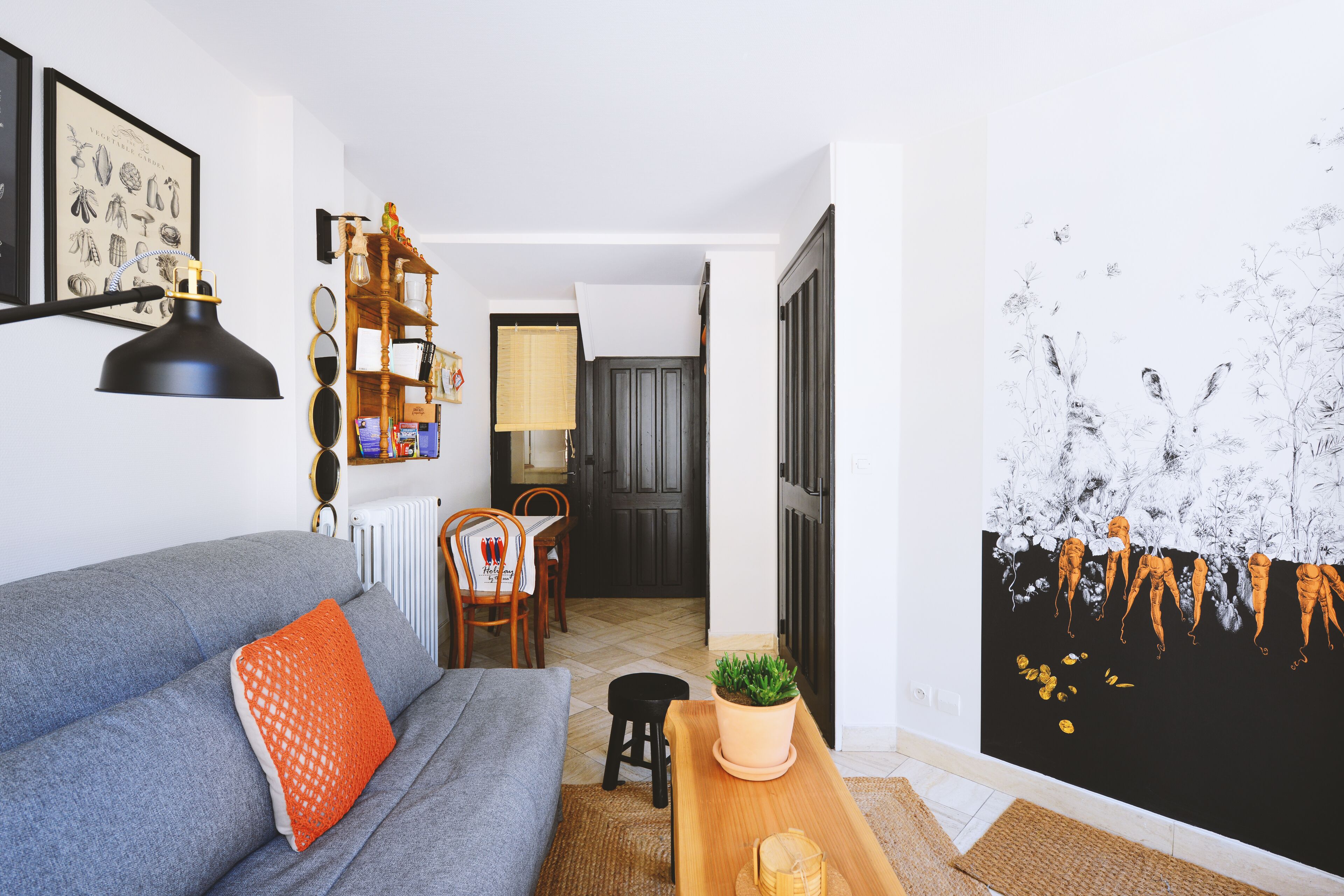 Un salon avec un canapé gris, un coussin orange, une table d’appoint en bois sur laquelle se trouve une plante, et une lampe noire.