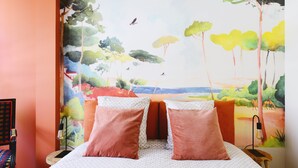 Une chambre à coucher avec un lit, deux oreillers, une table de chevet, une lampe et une fresque représentant un paysage sur le mur.
