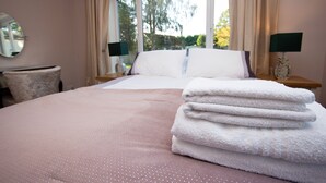 Een net opgemaakt bed met een roze sprei, een stapel witte handdoeken en een raam met gordijnen.