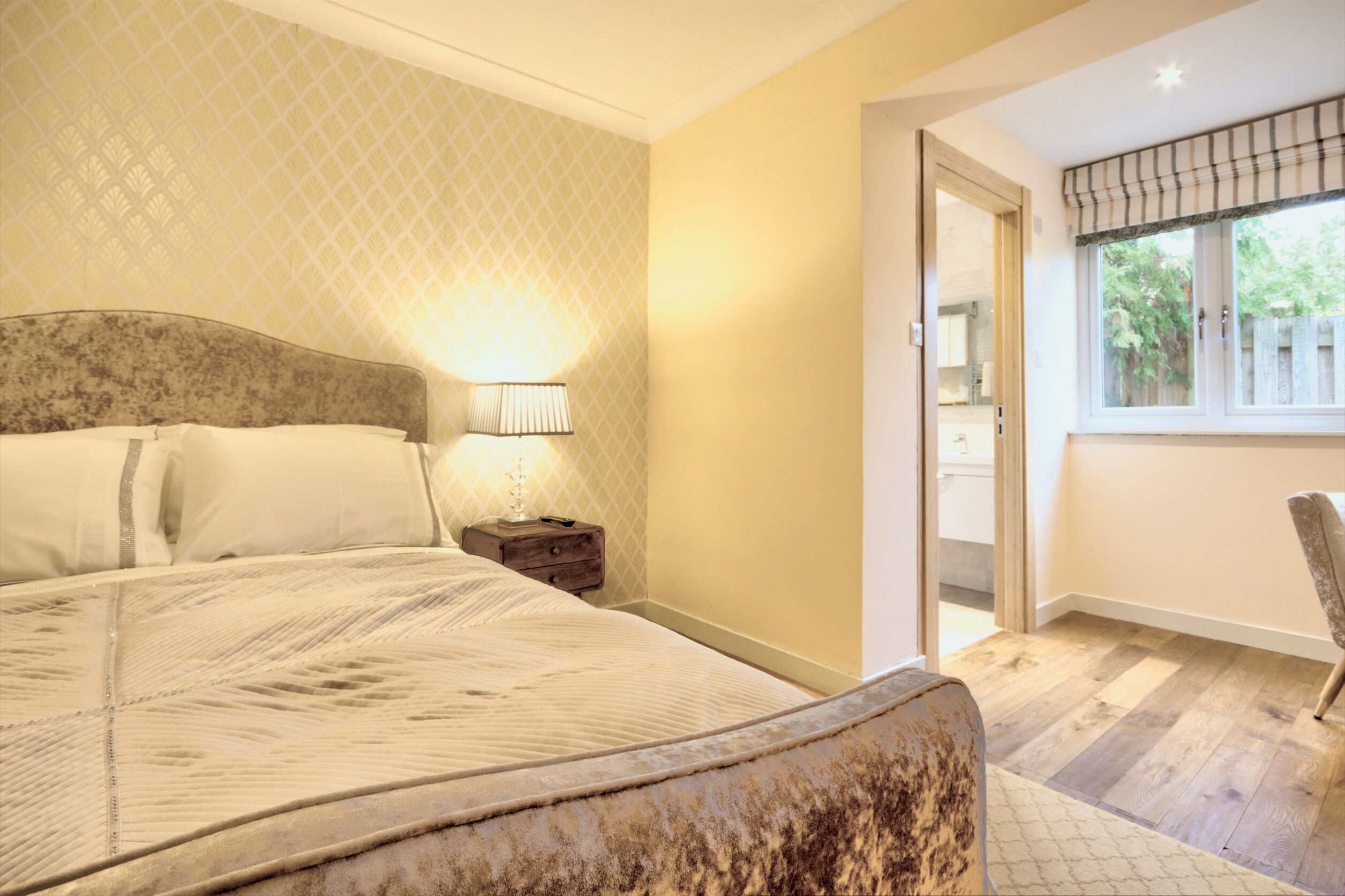 A bedroom with a large bed, a nightstand with a lamp, a chair, and a window with a view of the outside.