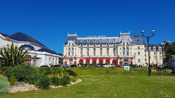 A grand hotel with a classic European architectural style, featuring a prominent entrance with red awnings and multiple flags. - Cocooning bubble and immersive veranda in a green setting 100m from the beach (Varaville)