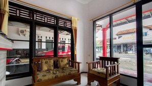 A room with a wooden sofa and chair, a large window with a view of a street and a building named "ELVANO". - OYO 3126 Penginapan Suka Asih (Pangandaran)