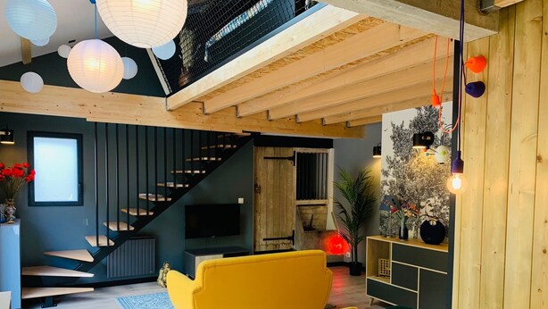 Een moderne woonkamer met een gele bank, houten balken in het plafond en een trap.