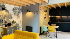 Een moderne woonkamer met een gele bank, een eethoek met een tafel en stoelen, en een keuken met donkere kasten en een magnetron.