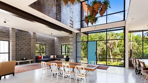 A spacious living area with a large dining table, white chairs, a sofa, and a view of greenery through large windows. - Makai House | Tranquil Folly Retreat (Folly Beach)