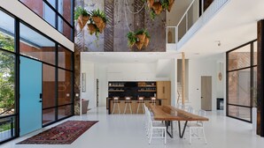 A spacious, modern interior with a long wooden dining table, white chairs, a kitchen area, and hanging planters. - Makai House | Tranquil Folly Retreat (Folly Beach)