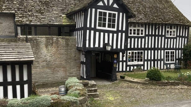 A traditional half-timbered building with a stone chimney, a small porch, and a gravel driveway.