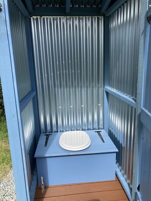 Een klein, afgesloten buitenterrastoilet met een metalen golfwand, een houten vloer en een witte wasbak.
