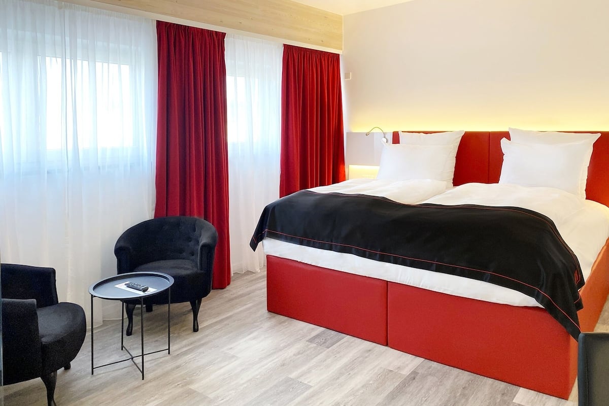 Habitación de hotel con una cama grande, cortinas rojas, una mesita con un control remoto y un sillón negro.