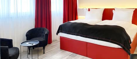 Habitación de hotel con una cama grande, cortinas rojas, una mesita con un control remoto y un sillón negro.