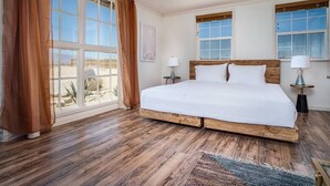 A bedroom with a large bed, wooden headboard, two bedside tables with lamps, and large windows with curtains.