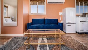 A living room with a blue sofa, a glass coffee table, a patterned rug, a white refrigerator, and a window with blinds.