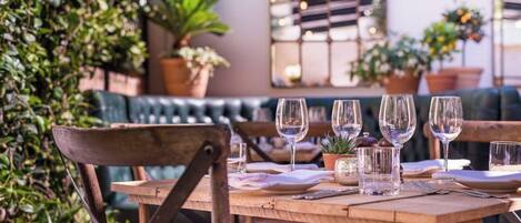 A wooden table set with wine glasses, plates, and cutlery, with a green plant and a window in the background.