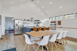 Una cocina moderna con una isla central, un comedor con mesa de madera y sillas blancas, y vistas al exterior a través de ventanas amplias.