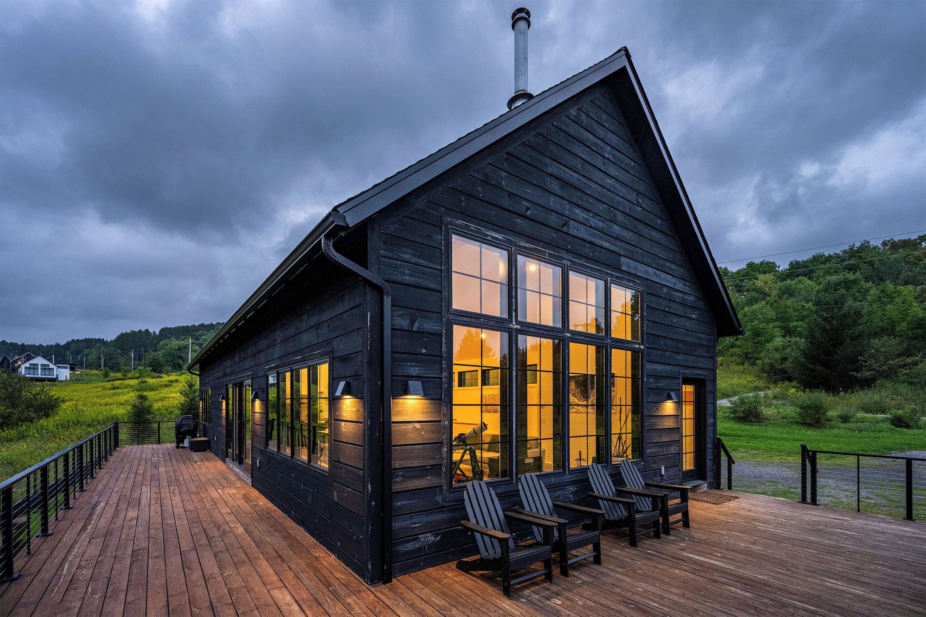 A wooden cabin with a deck and large windows, surrounded by greenery.