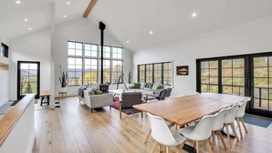 A spacious living area with a large wooden dining table, white chairs, a grey sofa, and a large window offering a view of the outdoors. - Magnificent Chalet, Sweeping Views and Hot Tub (Ellicottville)