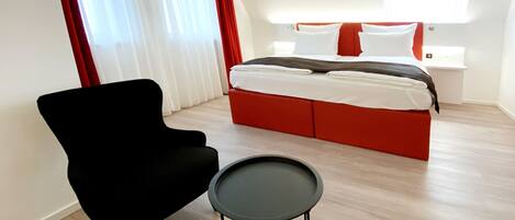 Una habitación de hotel moderna con una cama, un sillón negro, una mesita y un ventanal con cortinas rojas.