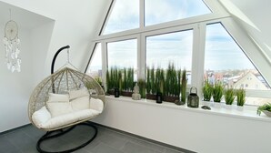 A modern room with a high ceiling, a hanging chair, a window with a view of the city, and a row of potted plants. - DORMERO Hotel Hersbruck (Hersbruck)