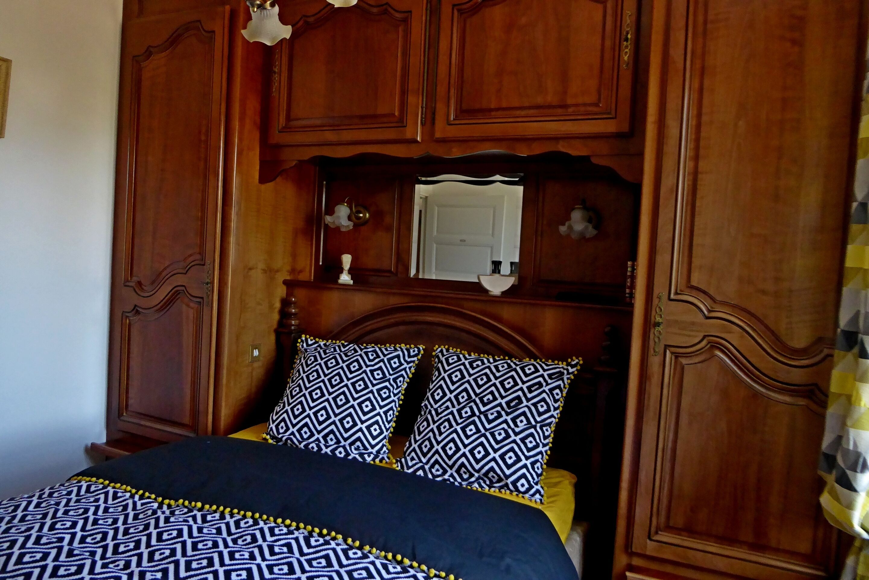 Ein hölzernes Bett mit einem gemusterten Bettbezug und zwei Kissen, ein Holz-Kopfstück und ein Spiegel darüber.
