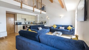 Een moderne woonkamer met blauwe banken, een keuken op de achtergrond en een houten trap.