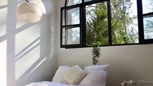 A bedroom with a bed, pillows, a bedside table with a plant, and a window with a view of trees. - DUPLEX Impressionnistes Barbizon Rive Gauche (Barbizon)