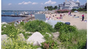 Ein Strand mit badenden Personen, ein Haus im Hintergrund und Boote im Wasser.