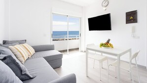 A modern living room with a grey sofa, a white dining table with chairs, a flat-screen TV, and a view of the sea through large windows.