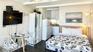 Une chambre d’hôtel comprenant un lit, un bureau avec une chaise, une télévision, un coin cuisine et un ventilateur de plafond.