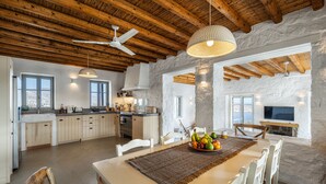 A spacious kitchen with a large island, white cabinetry, and a stone wall. There is a dining area with a table set for two, a fruit bowl, and a hanging light fixture. - Infinity Villa, luxury house with stunning view in Serifos (Serifos)