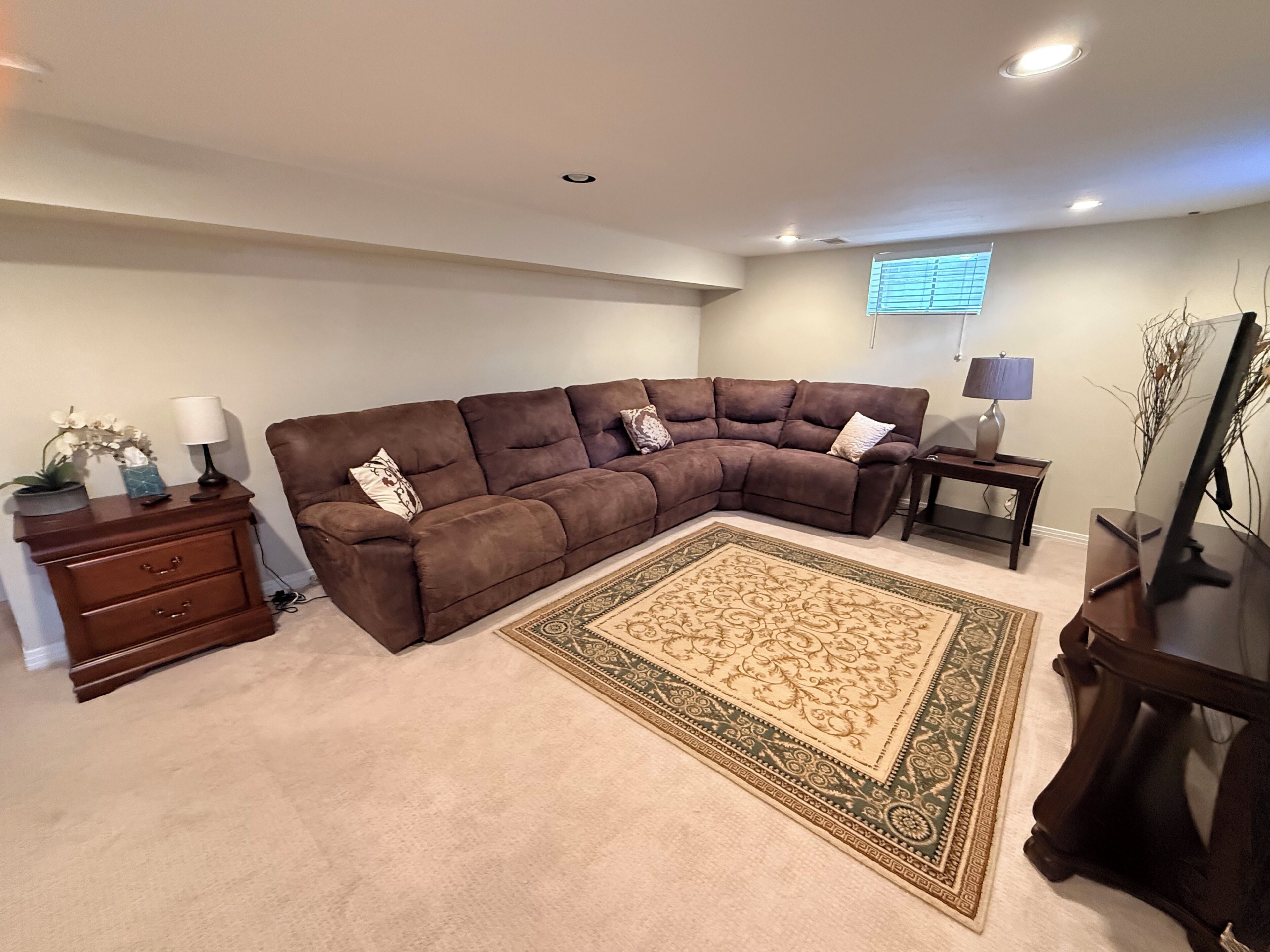 A living room with a brown sectional sofa, a wooden side table, a lamp, a desk, and a computer monitor.
