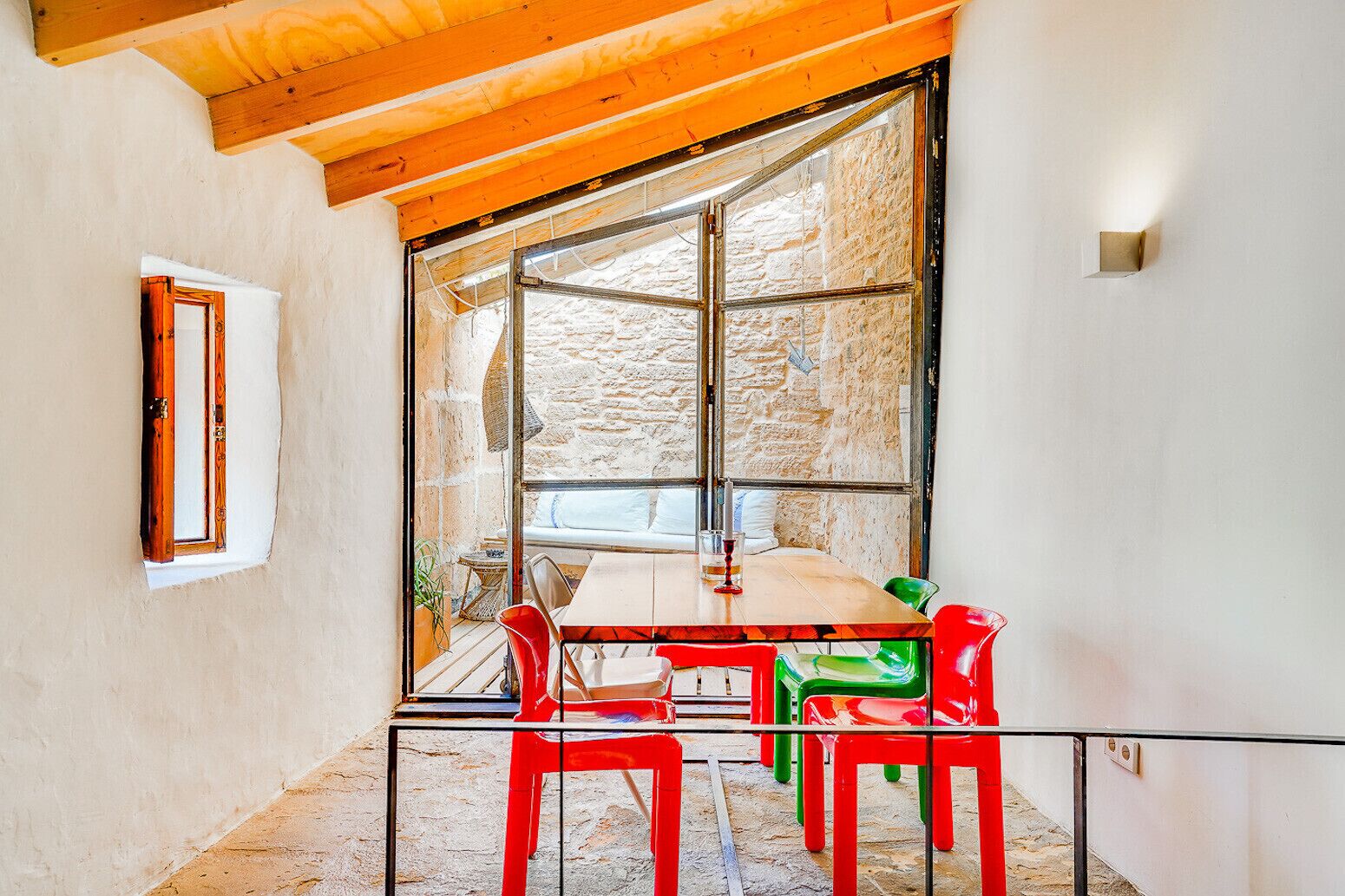 A dining area with a table and chairs, a window with a stone wall view, and a wooden ceiling.