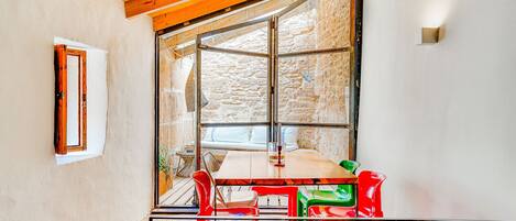 A dining area with a table and chairs, a window with a stone wall view, and a wooden ceiling.