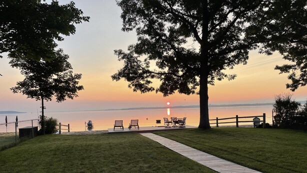 A grassy area with a wooden pathway leading to a seating area with chairs and a table, overlooking a calm body of water during sunset.