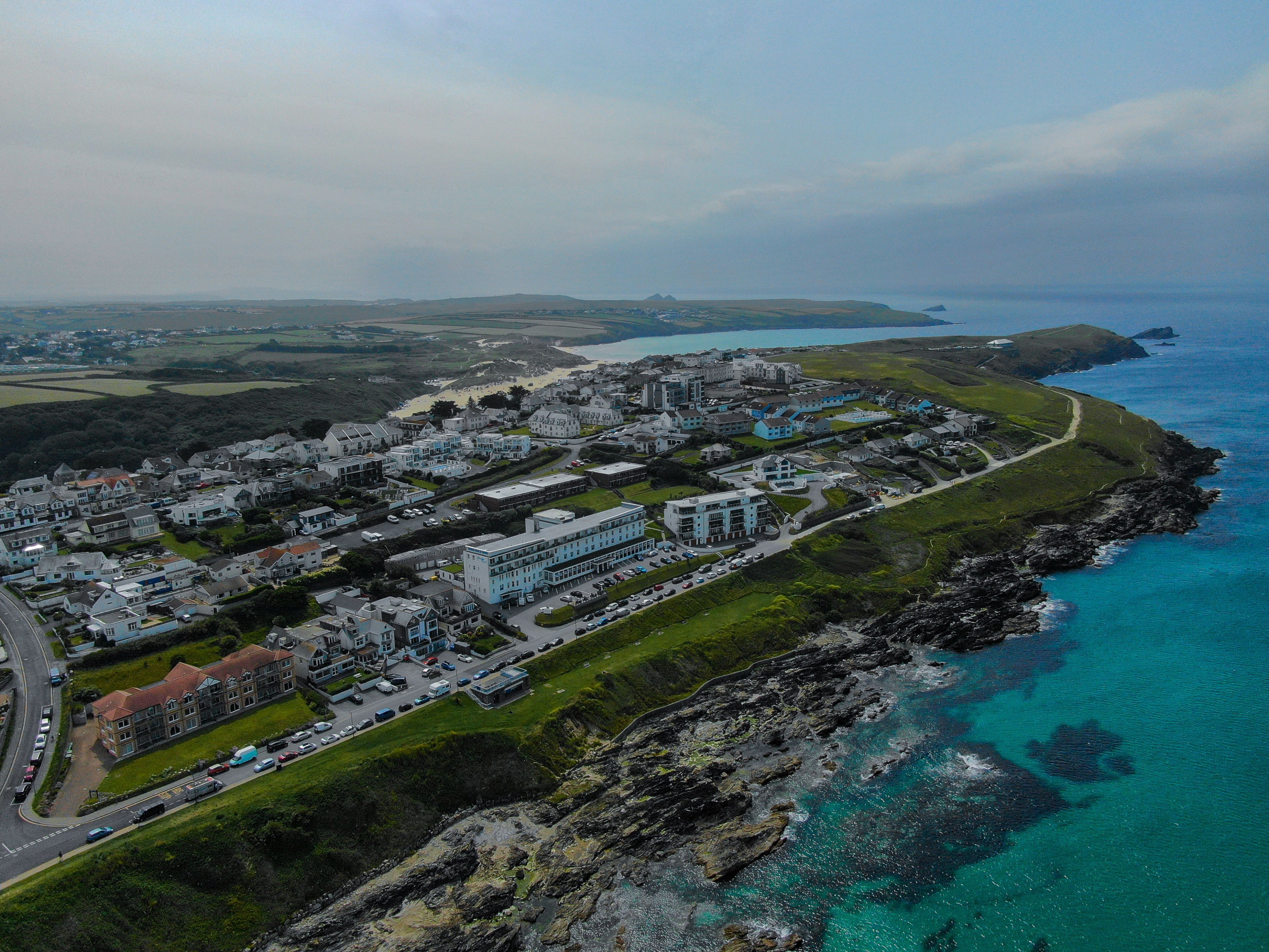 Vista aérea de un pueblo costero con una mezcla de edificios residenciales y comerciales, un hotel destacado con un amplio estacionamiento y un litoral rocoso.