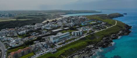 Vista aérea de un pueblo costero con una mezcla de edificios residenciales y comerciales, un hotel destacado con un amplio estacionamiento y un litoral rocoso.