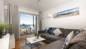 A modern living room with a grey sofa, a coffee table, and a view of the sea through a glass door. - Fistral Lookout - Parking, WiFi & Sea View (Newquay)
