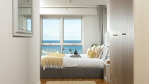 A bedroom with a bed, a wardrobe, a window with a view, and a bedside table with a plant. - Fistral Lookout - Parking, WiFi & Sea View (Newquay)