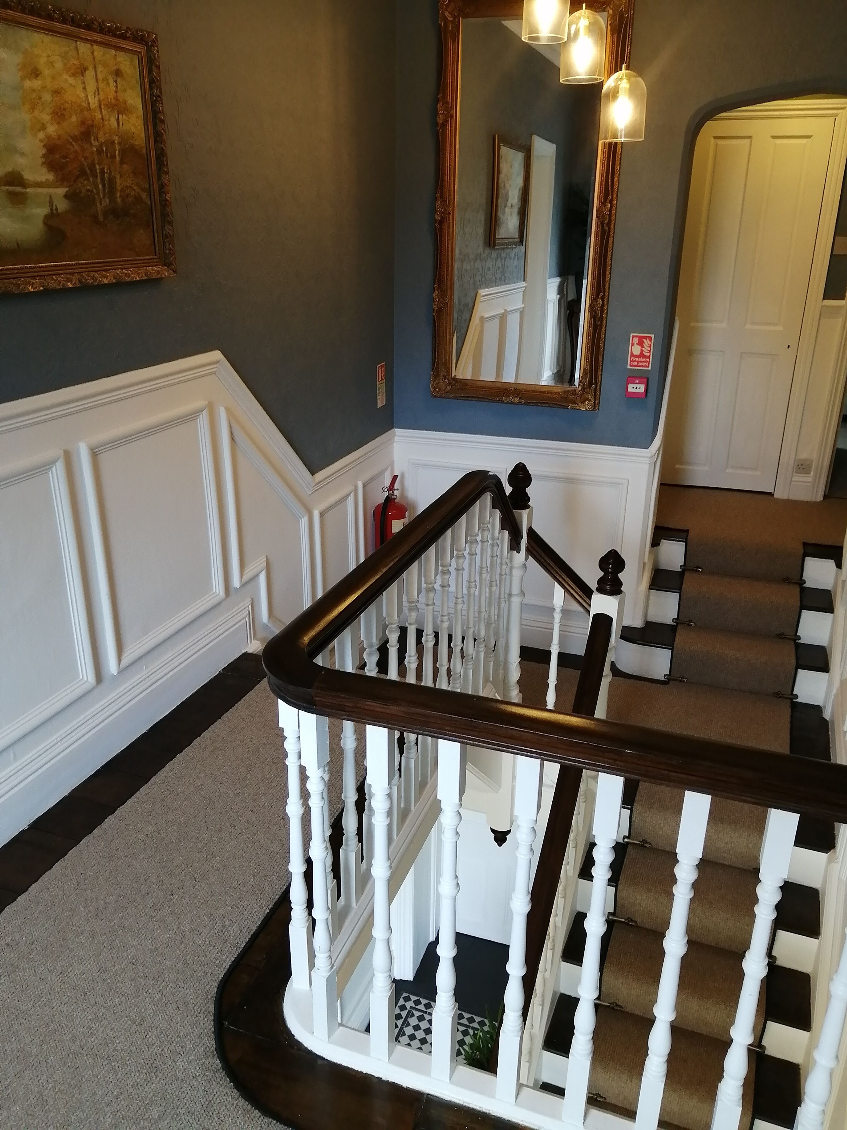 A staircase with white balusters and dark wooden handrails, leading to a landing with a large mirror and a framed painting on the wall.