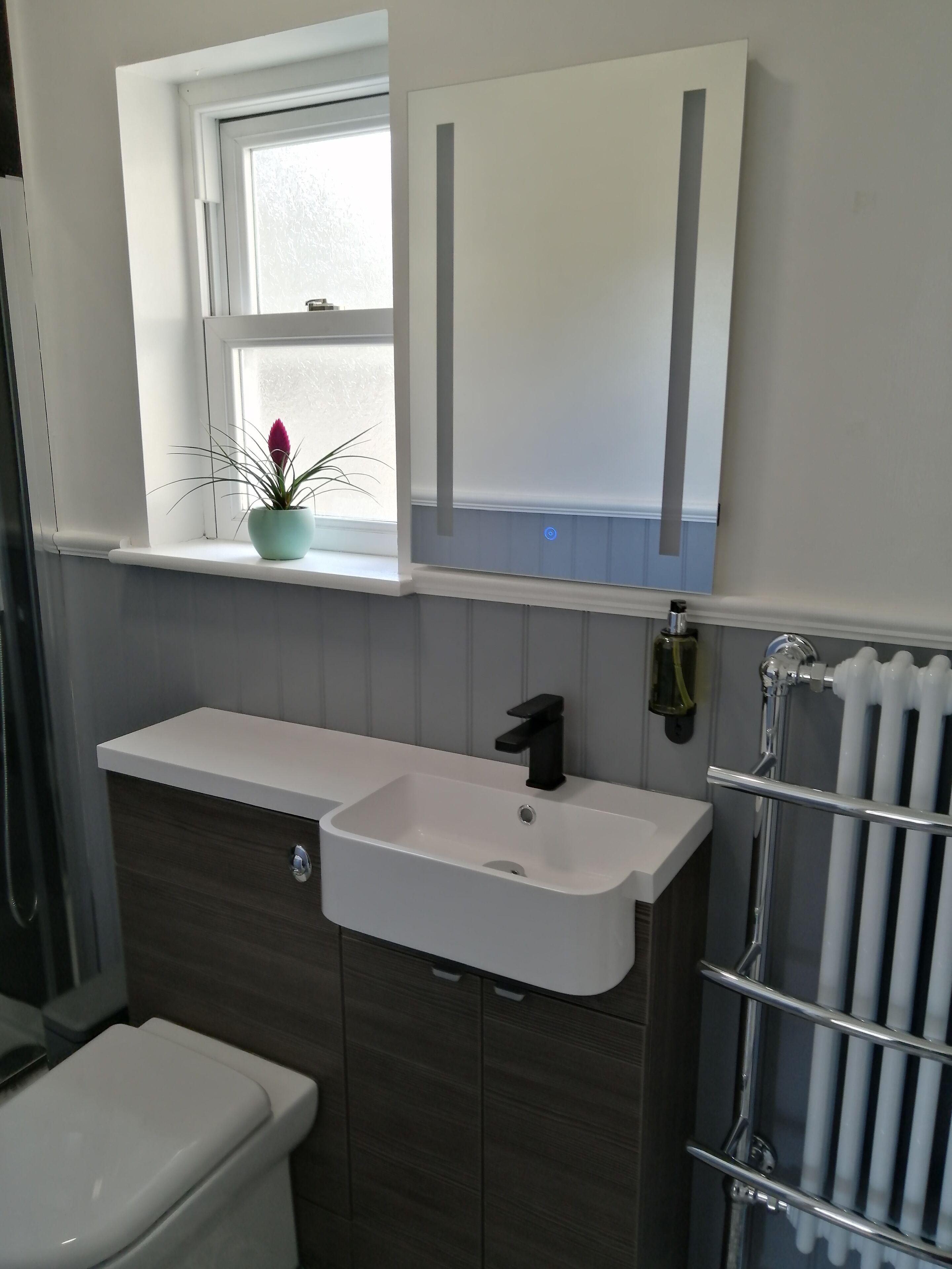 Un baño con lavamanos blanco, mueble de madera oscura, una ventana con planta y un inodoro blanco.