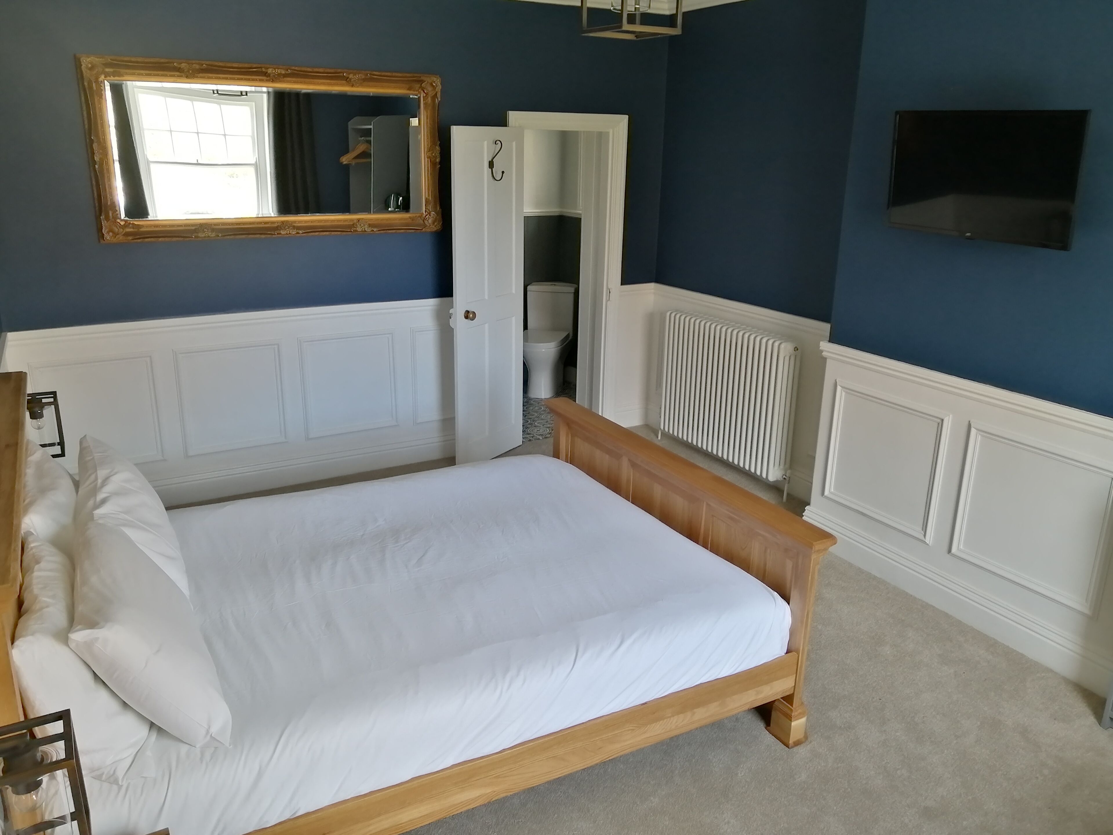 A bedroom with a wooden bed, white bedding, a large mirror, a white radiator, and a wardrobe.