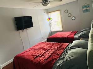 Ein Schlafzimmer mit zwei Betten, einem Deckenventilator, einem an der Wand montierten Fernseher und einem Fenster mit Jalousien.