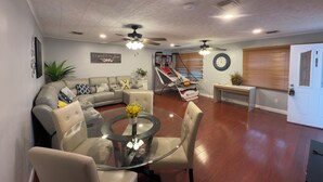 A living room with a sectional sofa, a glass coffee table, a dining table with chairs, a ceiling fan, and a basketball hoop.