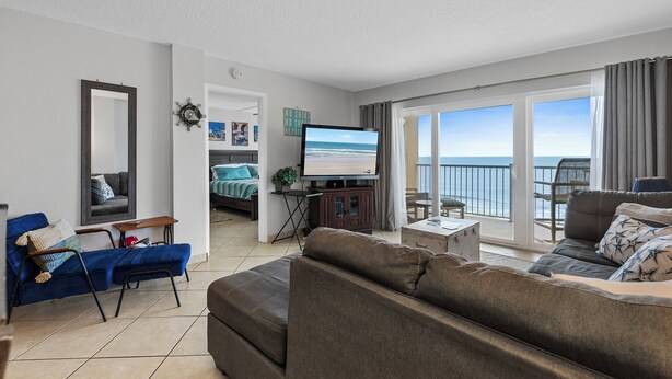 A modern living room with a large flat-screen TV, a sofa, a blue armchair, and a balcony with a view of the sea. - Direct Oceanfront View from Nearly Every Room! Great Getaway Near No-Drive Beach (New Smyrna Beach)