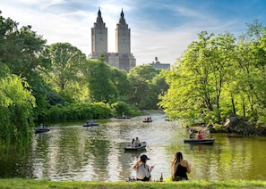 A river with boats, surrounded by lush greenery and two prominent buildings in the background. - Nap York Central Park Sleep Station (New York)