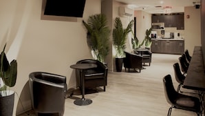 Un moderno vestíbulo de hotel con un televisor de pantalla plana montado en la pared, un rincón de descanso con sillas y mesas negras, y una zona de cocina con armarios oscuros y un microondas.